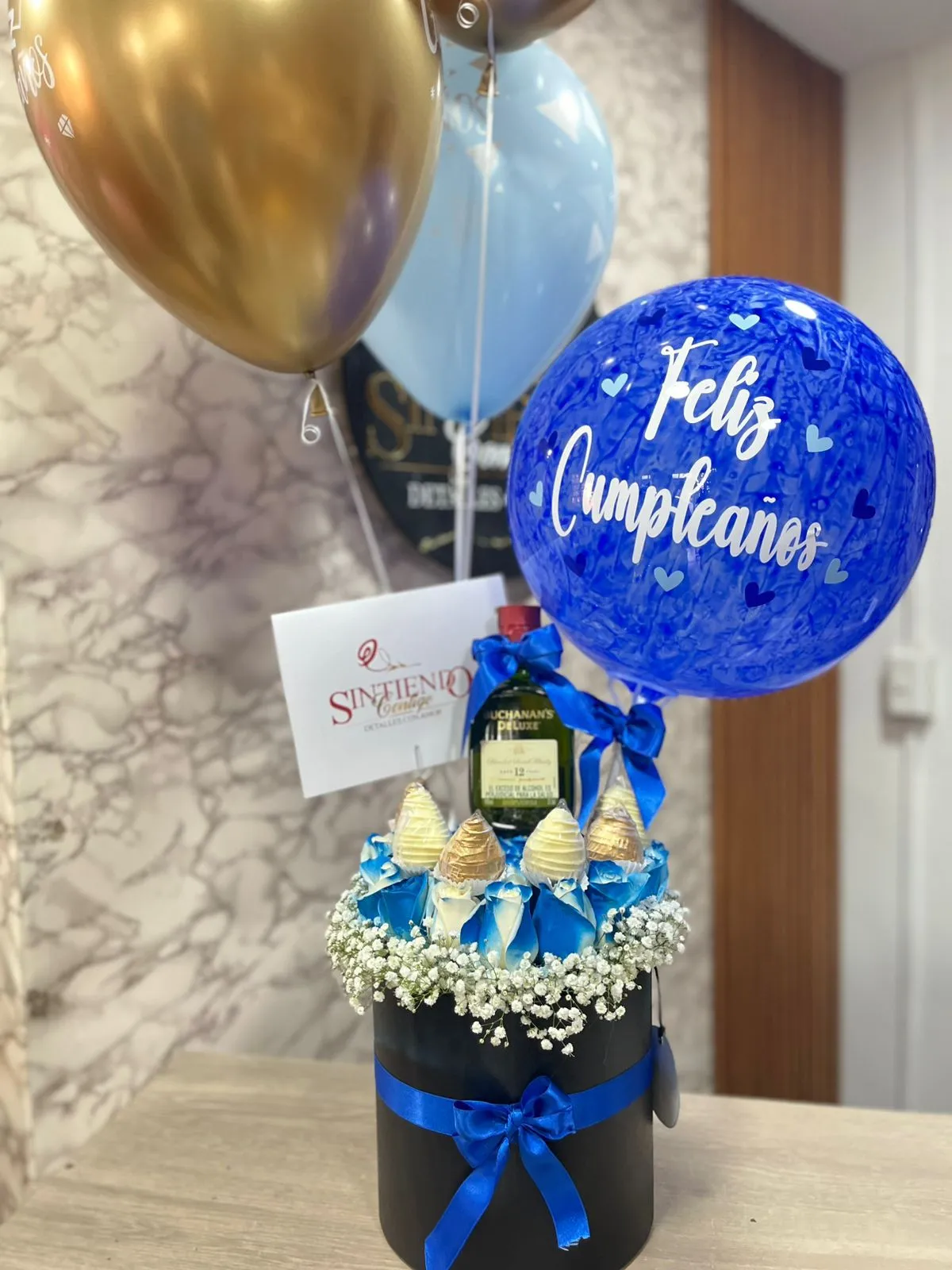Cilindro con rosas azules, fresas con chocolate, botella de Buchanas y globos con helio